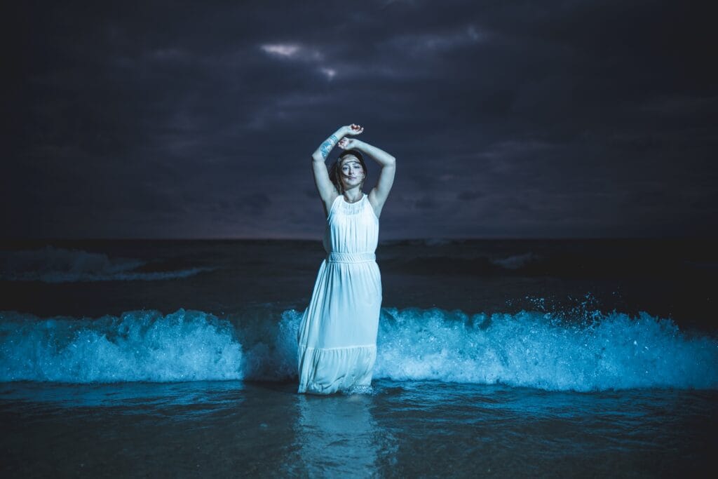 Kaitlyn in a flowing white dress in blue surf beneath storm clouds.