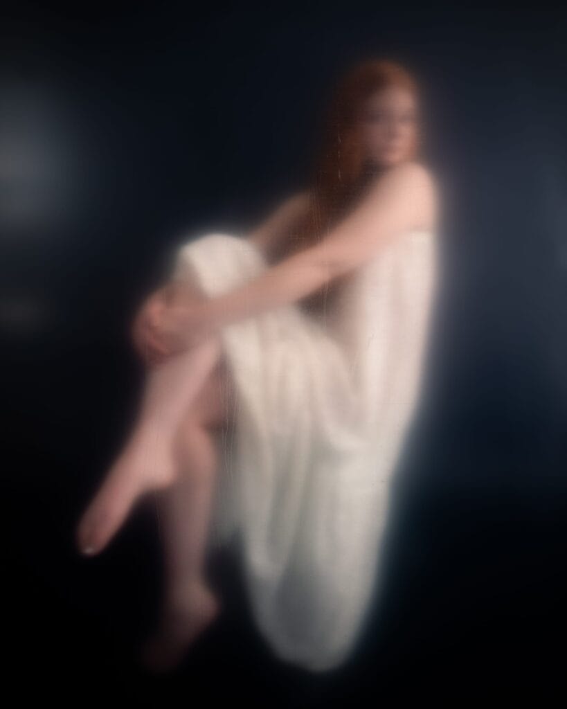 Elizabeth sits with her legs crossed, photographed through a softly obscuring surface.