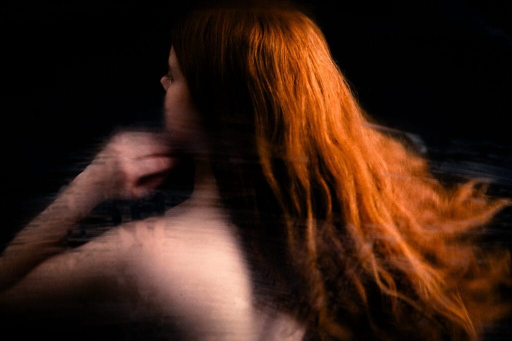 Elizabeth turns away, her red hair flowing across a dark portrait with horizontal optical distortion.