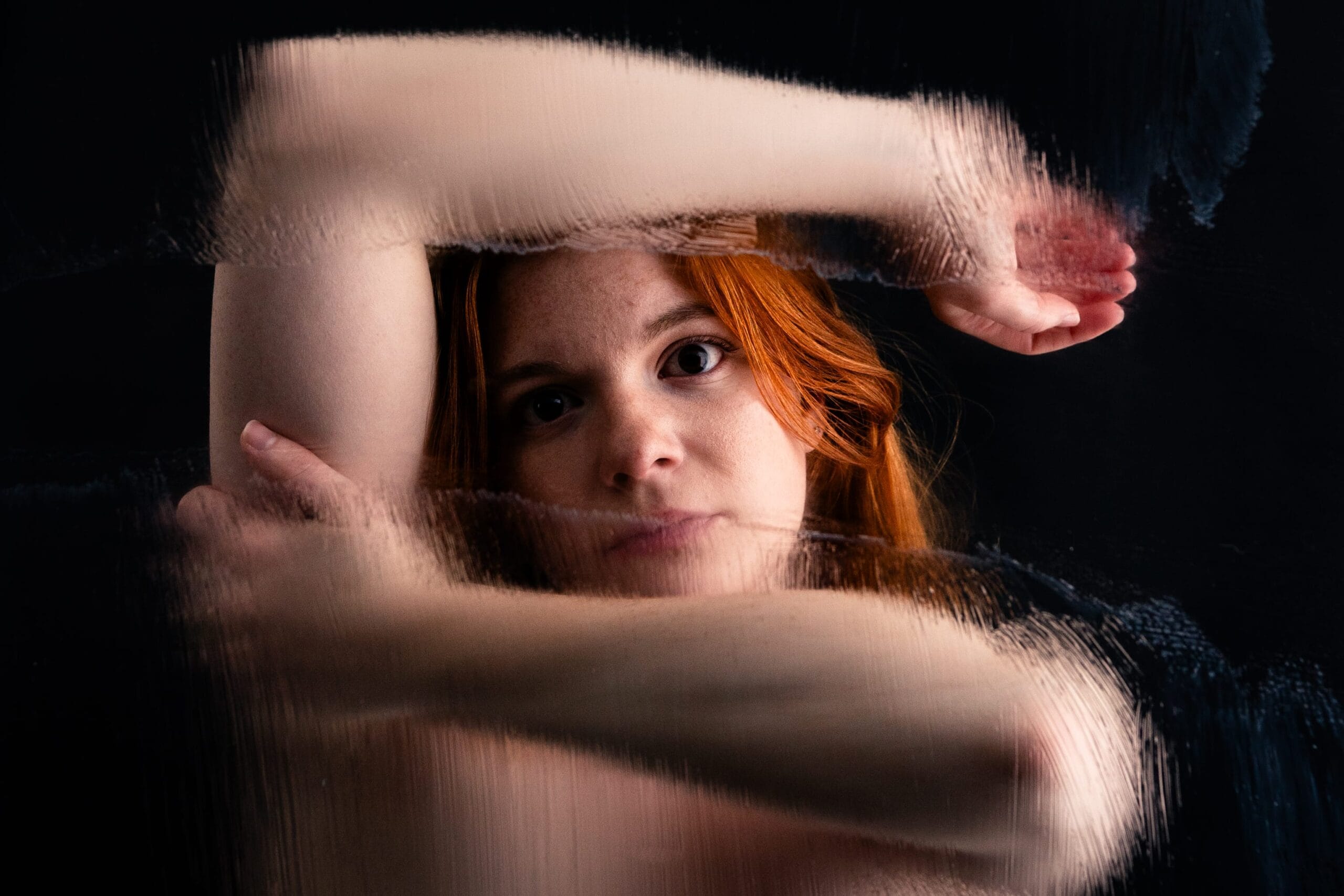 Elizabeth frames her face with raised arms behind a surface that blurs and warps their outlines.