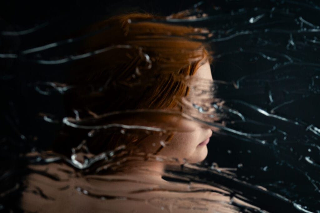 Elizabeth in profile with red hair, photographed through a streaked surface that distorts her face.