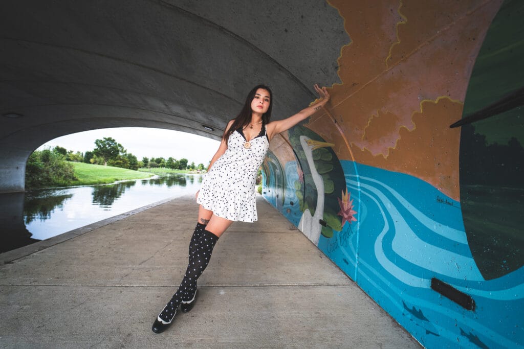 Reese leans against a colorful mural beside the river beneath a concrete bridge.