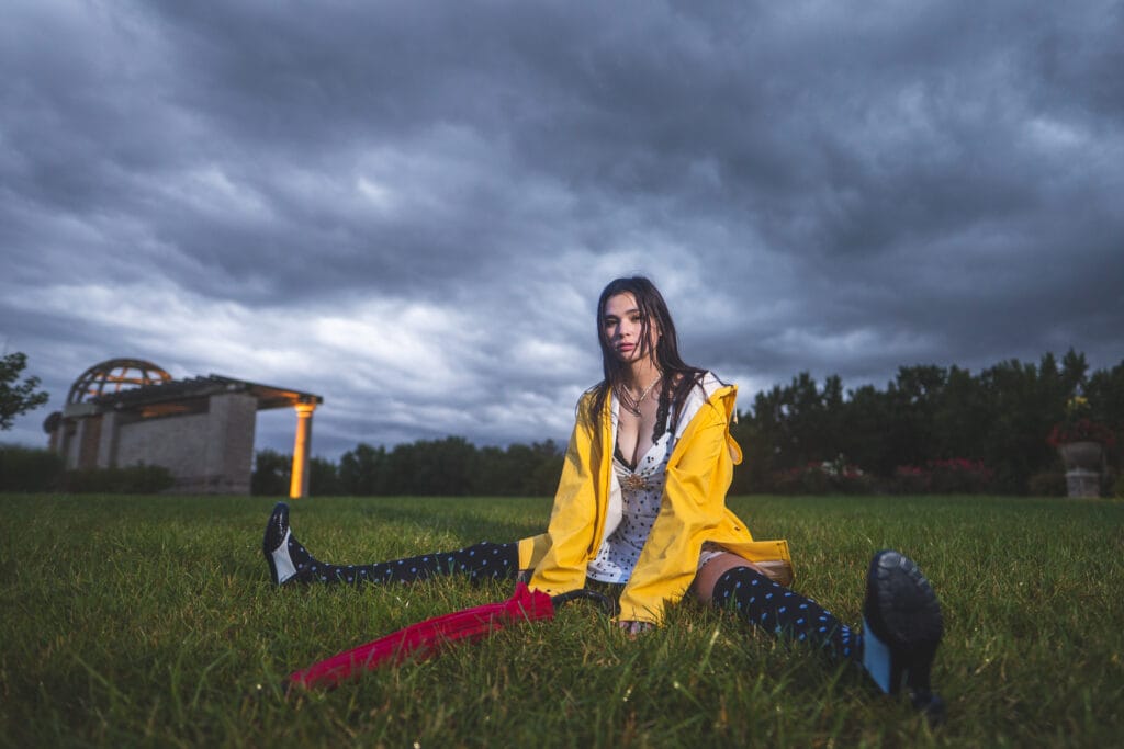 Reese sits on wet grass with a red umbrella and yellow raincoat beneath dark clouds.