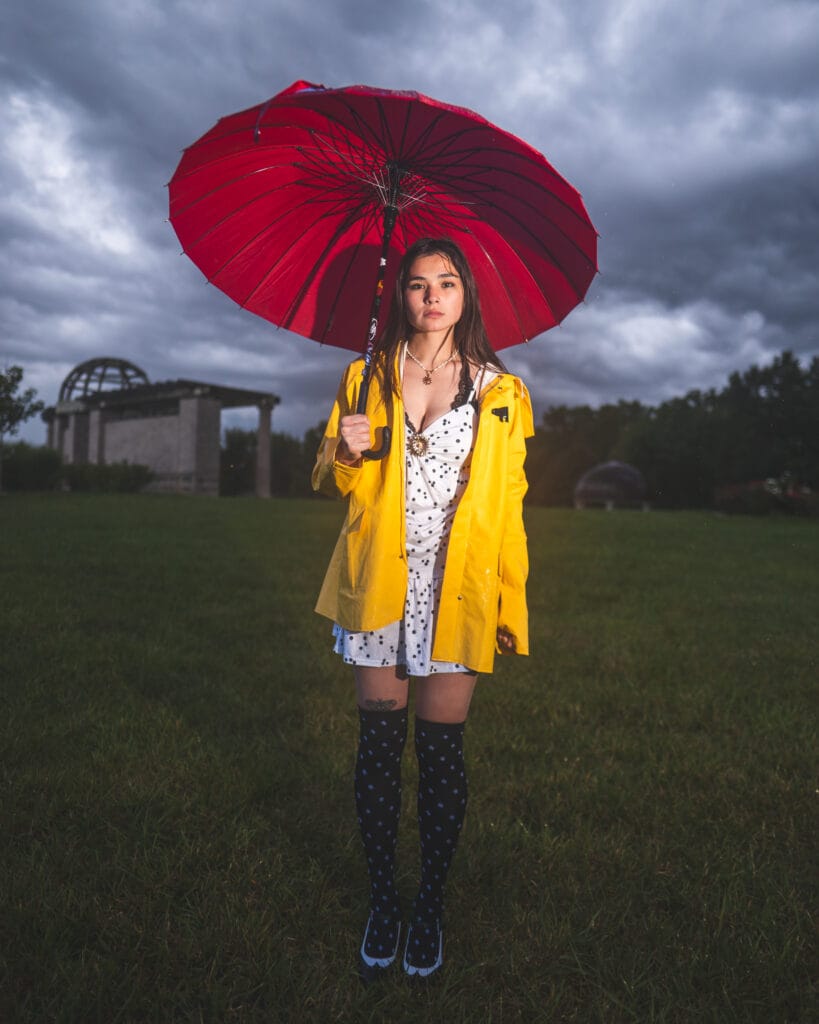 Reese holds a red umbrella while wearing a yellow raincoat beneath storm clouds.