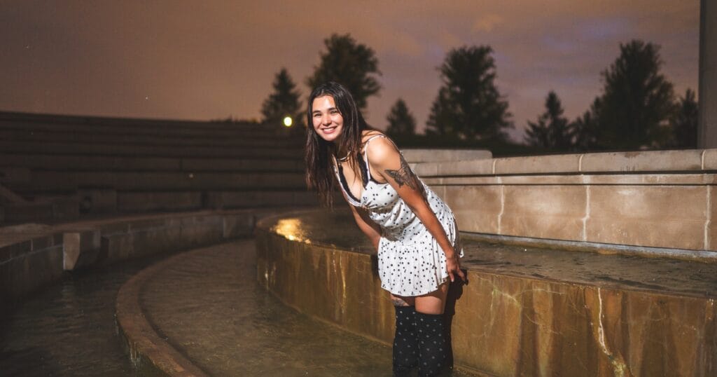 Reese smiles beside a curved fountain in the evening, wearing a white polka-dot dress.