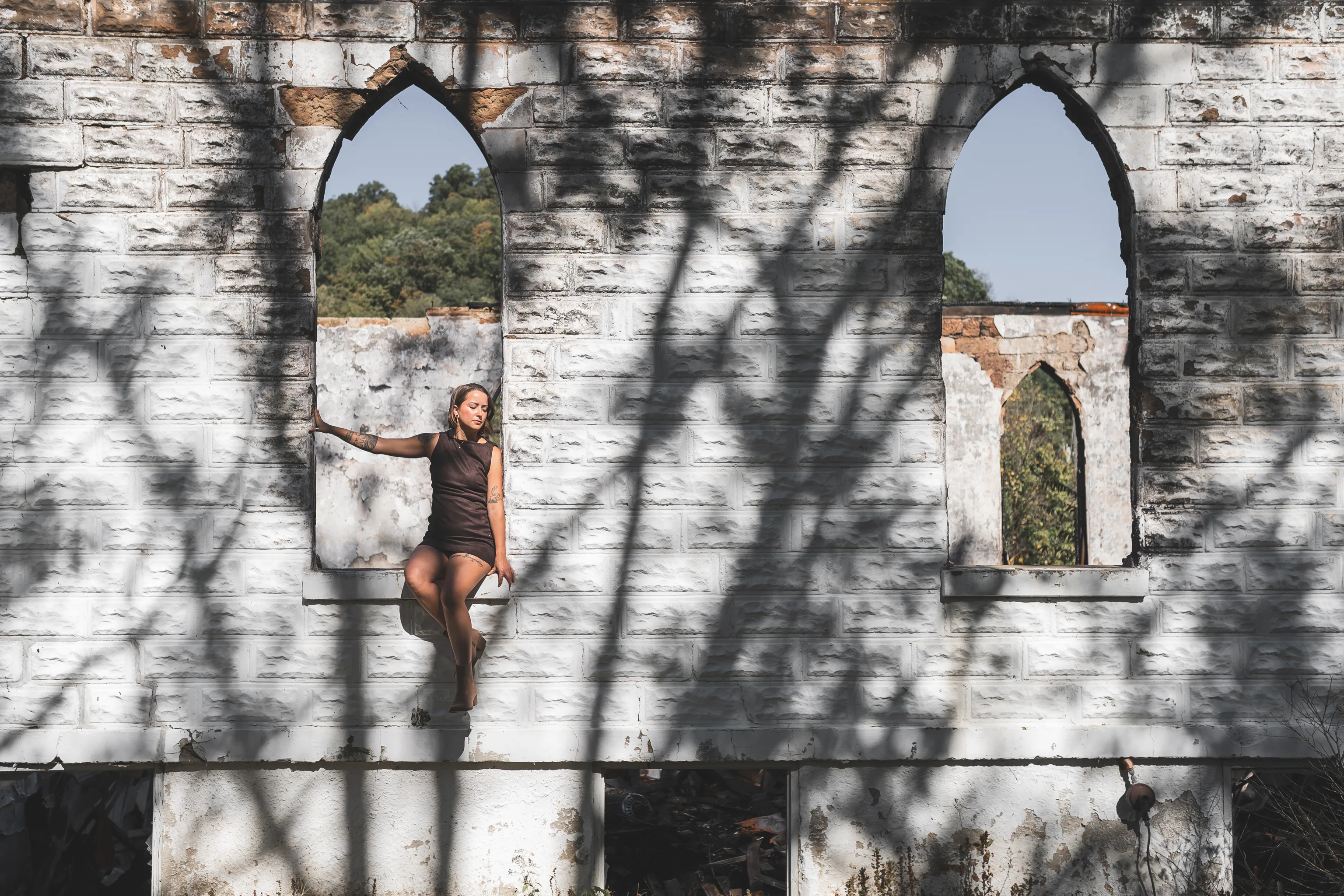 Kaitlyn sits in an empty pointed window in her brown dress, surrounded by white stone walls and branching shadows.