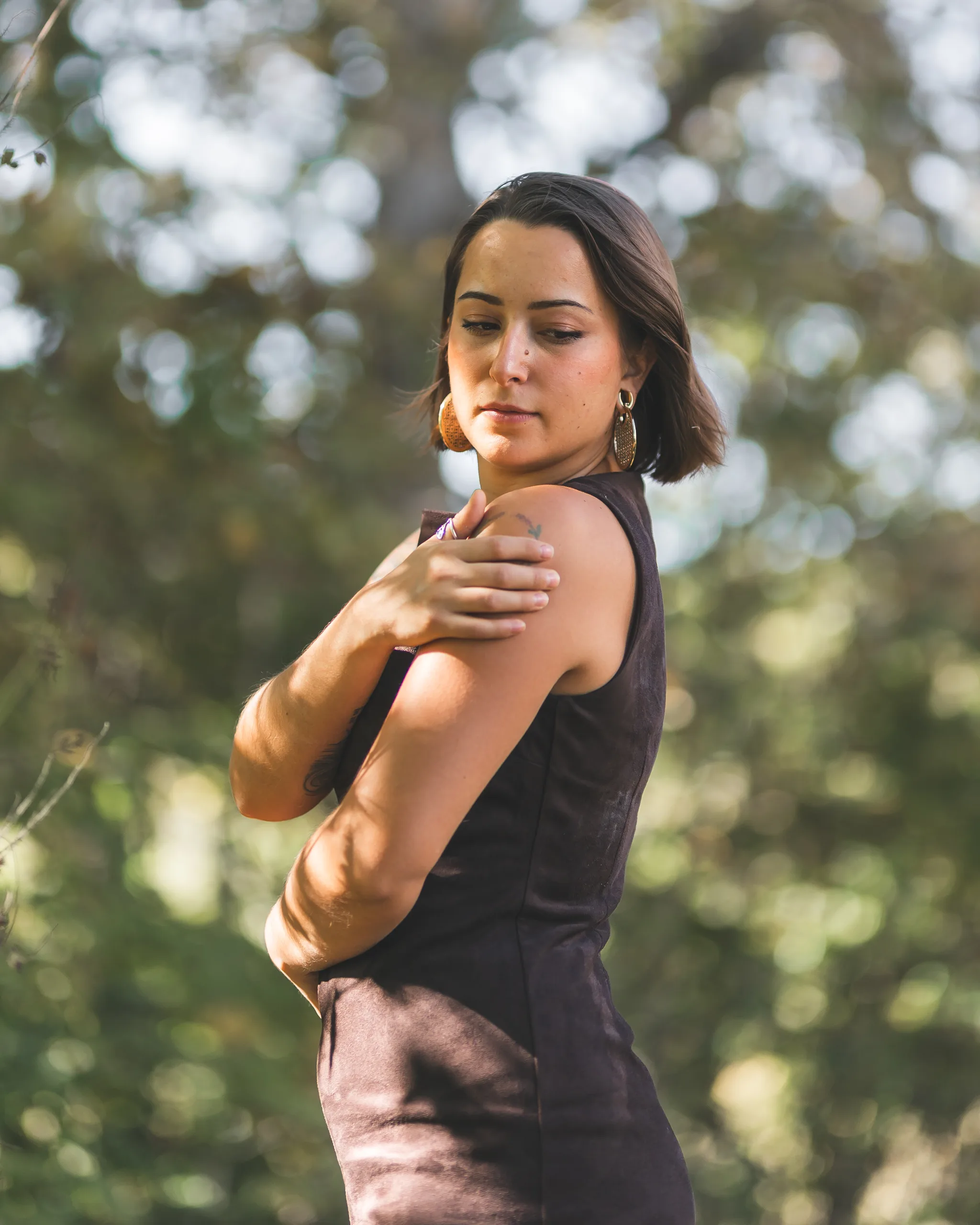 Kaitlyn rests one hand on her shoulder in her brown dress beneath dappled tree light.