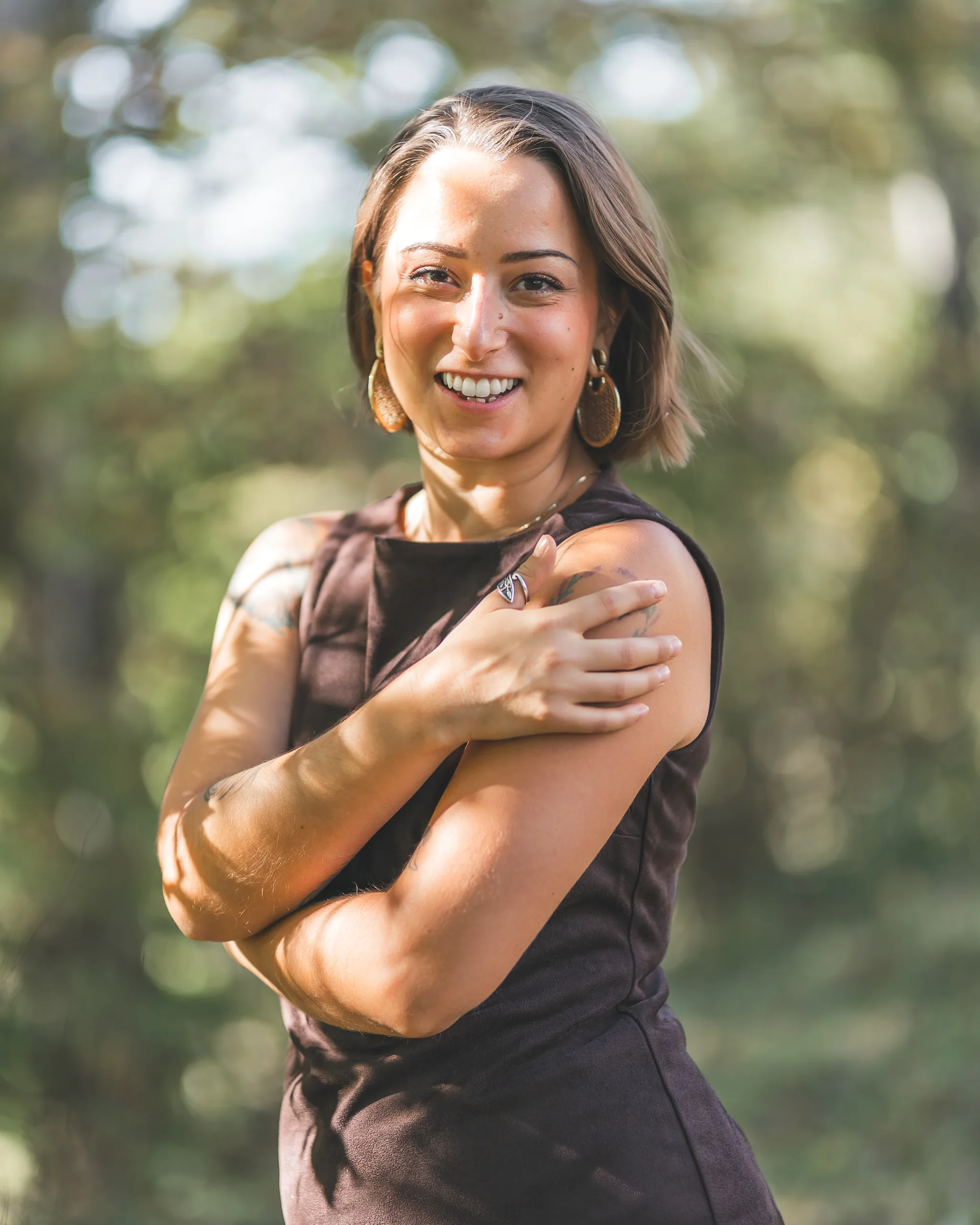 Kaitlyn laughs beneath the trees in her brown dress, with sunlight falling across her face.