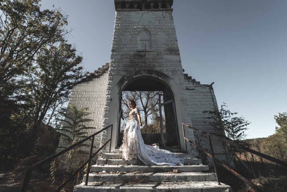 Kaitlyn stands on the steps of a burned church in a distressed vintage wedding dress, its long train spread behind her.