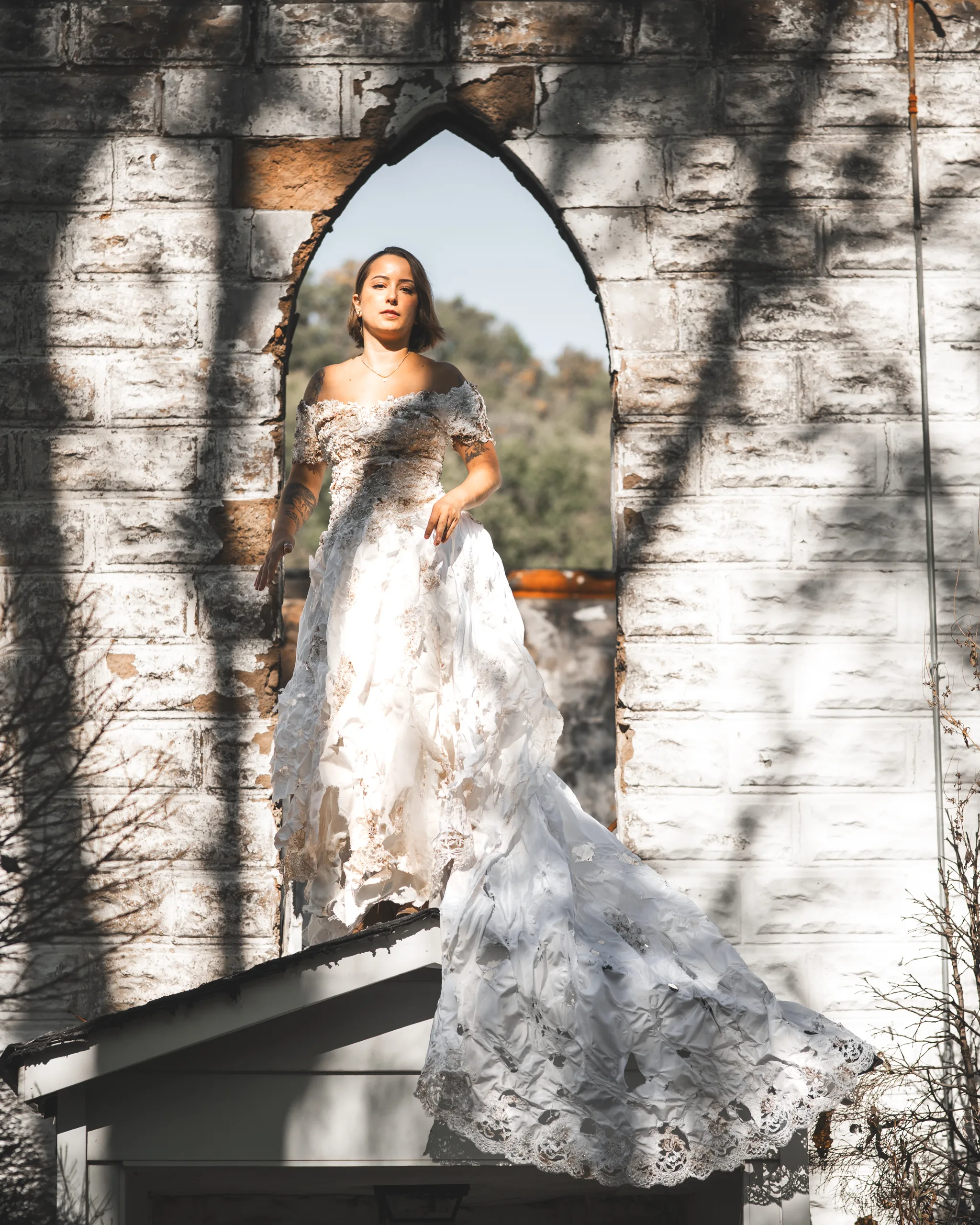 Kaitlyn stands in a pointed church window wearing the distressed vintage wedding dress, with its train draped across a beam.