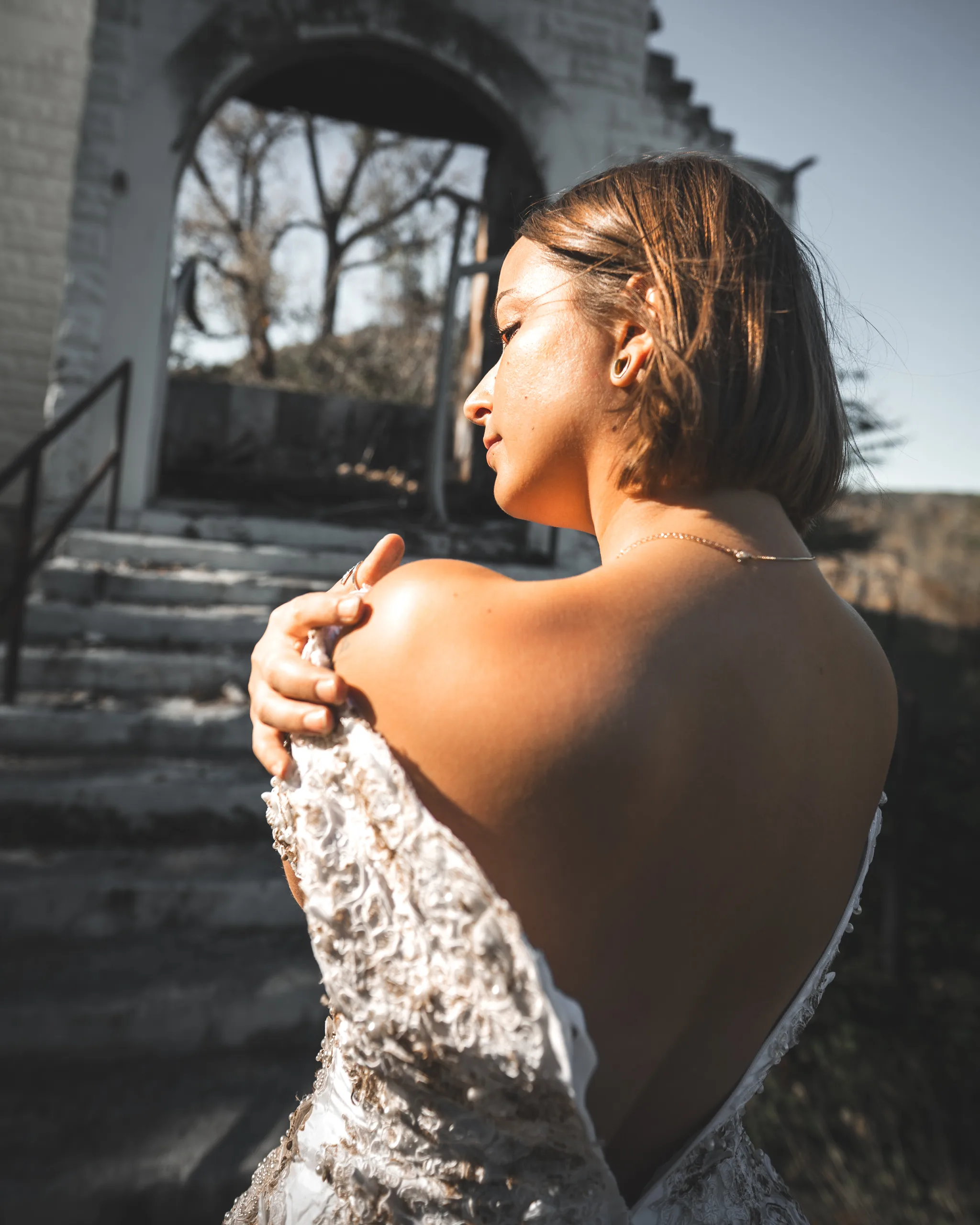 Kaitlyn looks back over her bare shoulder, holding the wedding dress against her chest near the church steps.